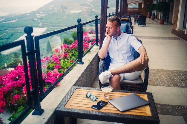 Young handsome businessman in a white shirt and shorts sits with a laptop in a cafe at the table Work while resting