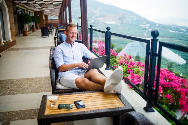 Young handsome businessman in a white shirt and shorts sits with a laptop in a cafe at the table. Work while resting