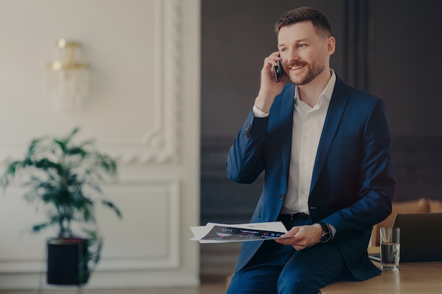 Young handsome businessman working from home, male entrepreneur in formal suit talking on phone in office, smiling discussing business ideas with copartner, having laptop and glass of water on desk