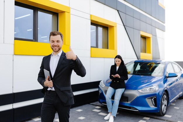 Young handsome car dealership consultant holding tablet while standing near car and showing finger up gesture