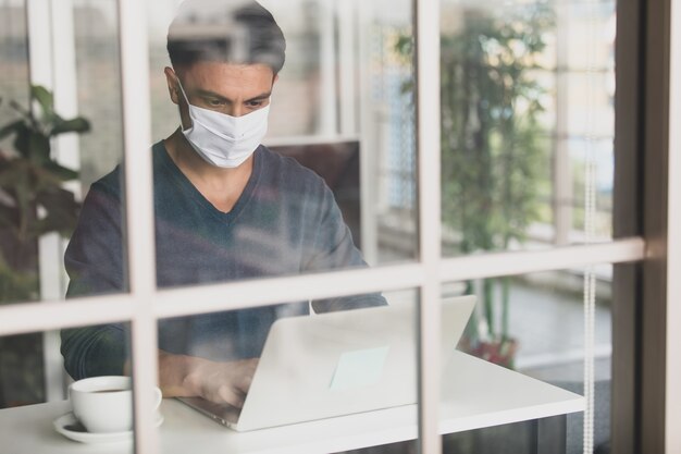 Young and handsome Caucasian businessman wearing medical hygiene protective mask sitting in house, drinking coffee and using laptop notebook computer. Idea for working from home in coronavirus crisis.