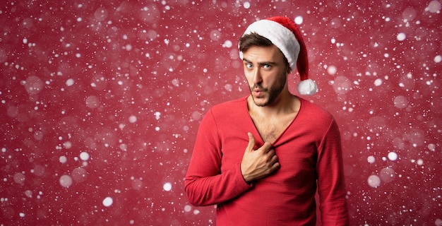 Young handsome caucasian guy in Santa hats stands on red background in studio pulls the sweater Shows how hot he is. Close up portrait european guy with Christmas mood. Holiday banner. New Year.