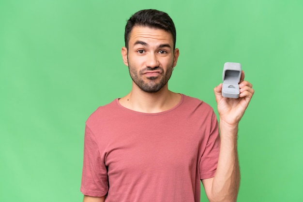 Young handsome caucasian man holding a engagement ring over isolated background with sad expression