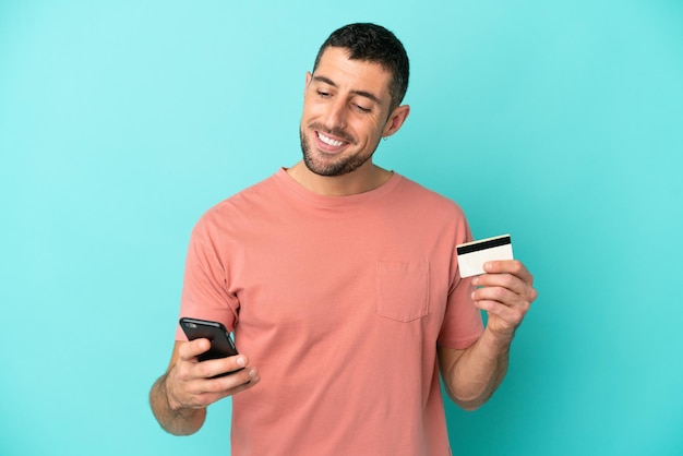 Young handsome caucasian man isolated on blue background buying with the mobile with a credit card