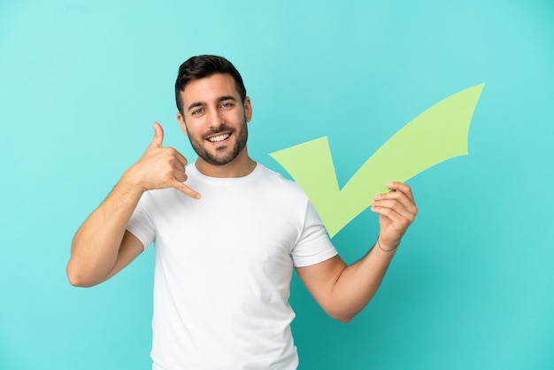 Young handsome caucasian man isolated on blue background holding a check icon and doing phone gesture