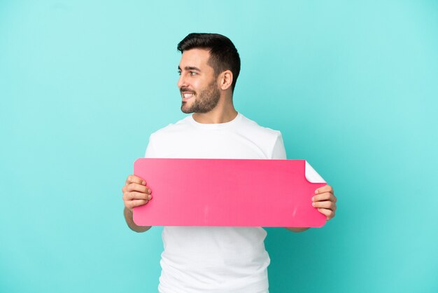 Photo young handsome caucasian man isolated on blue background holding an empty placard and looking side