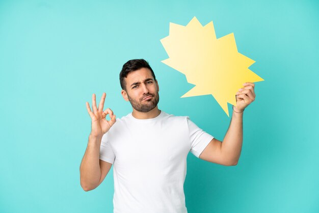 Young handsome caucasian man isolated on blue background holding an empty speech bubble and doing OK sign