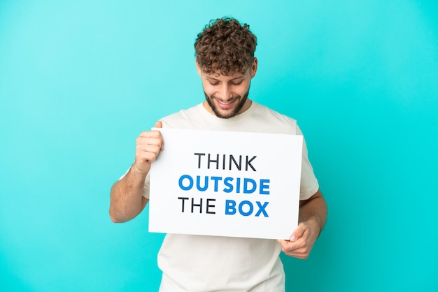 Young handsome caucasian man isolated on blue background holding a placard with text Think Outside The Box