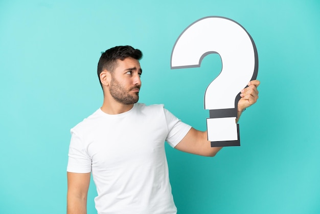 Young handsome caucasian man isolated on blue background holding a question mark icon and having doubts