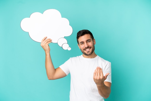 Young handsome caucasian man isolated on blue background holding a thinking speech bubble and doing coming gesture