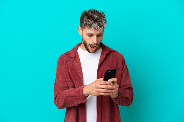 Photo young handsome caucasian man isolated on blue background looking at the camera while using the mobile with surprised expression