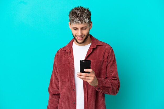 Photo young handsome caucasian man isolated on blue background sending a message or email with the mobile