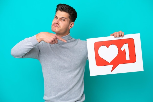 Young handsome caucasian man isolated on blue bakcground holding a placard with Like icon and pointing it