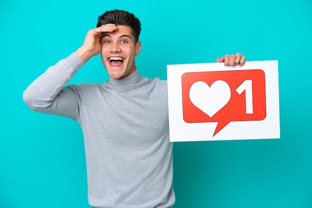 Young handsome caucasian man isolated on blue bakcground holding a placard with Like icon with surprised expression