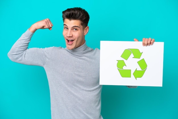 Young handsome caucasian man isolated on blue bakcground holding a placard with recycle icon and doing strong gesture