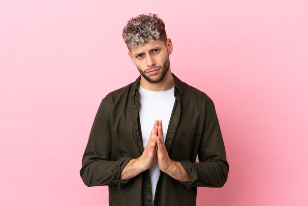 Young handsome caucasian man isolated on pink background keeps palm together. Person asks for something