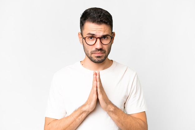 Young handsome caucasian man over isolated white background keeps palm together Person asks for something