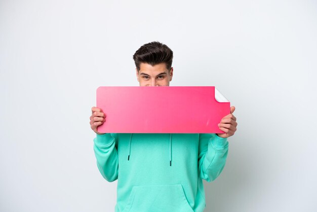 Photo young handsome caucasian man isolated on white bakcground holding an empty placard and hiding behind it