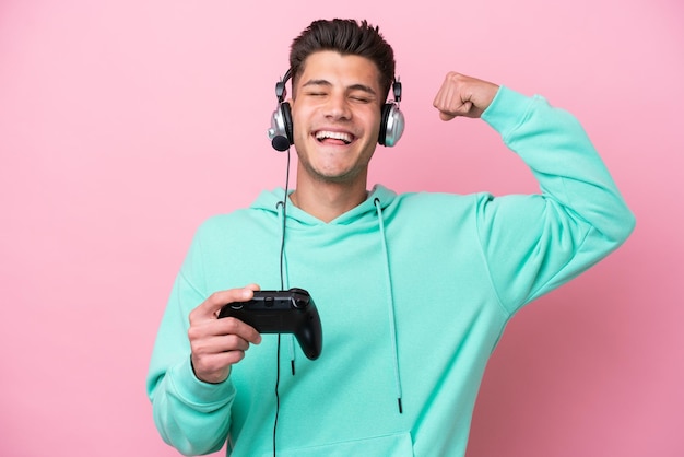 Young handsome caucasian man playing with a video game controller isolated on pink background doing strong gesture