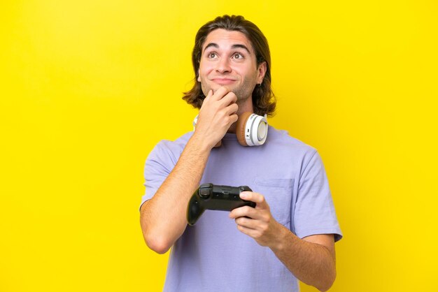 Young handsome caucasian man playing with a video game controller over isolated on yellow background having doubts and thinking