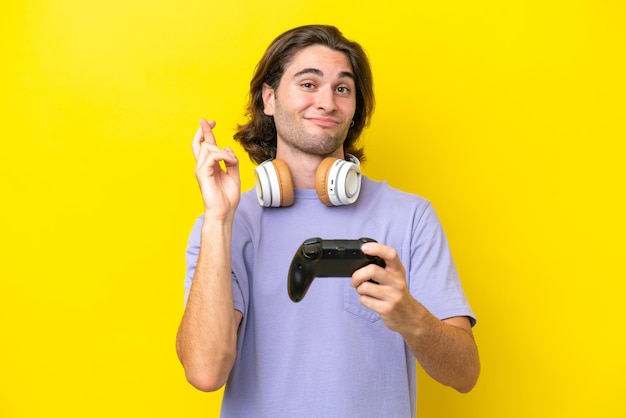 Young handsome caucasian man playing with a video game controller over isolated on yellow background with fingers crossing and wishing the best