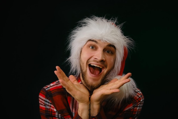 Young handsome Caucasian man in red fluffy Santa Claus hat on dark studio background