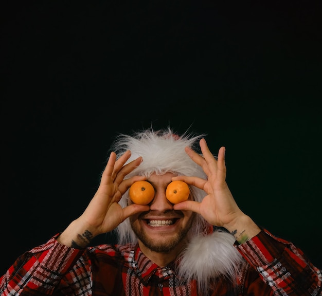 Young handsome Caucasian man in red fluffy Santa Claus hat holds tangerines with both hands