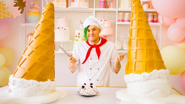 Young handsome chef with green dreadlocks with cake on table holding knife looking at camera smiling spreading hands standing in bright cafeteria