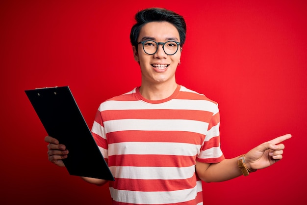 Young handsome chinese inspector man wearing glasses holding clipboard checklist very happy pointing with hand and finger to the side