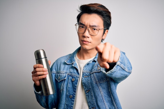 Young handsome chinese tourist man wearing glasses holding thermo with water pointing with finger to the camera and to you hand sign positive and confident gesture from the front