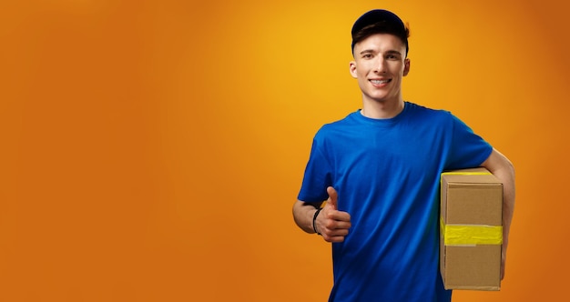 Young handsome delivery man holding parcel cardboard box against yellow background