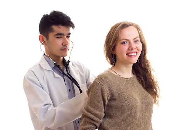 Young handsome doctor in white gown with stethoscope listening to the back of young pretty woman