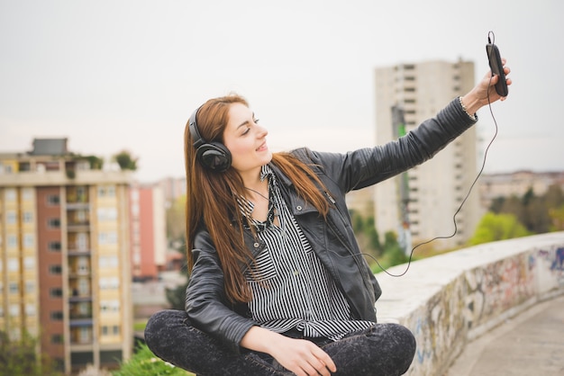 Young handsome eastern brunette girl listening music sitting on a small wall with city in background