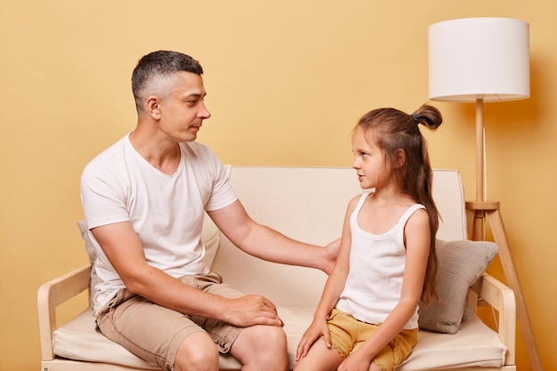 Young handsome father talking with his daughter sitting on cough at home against beige wall having conversation about their relationship looking at each other