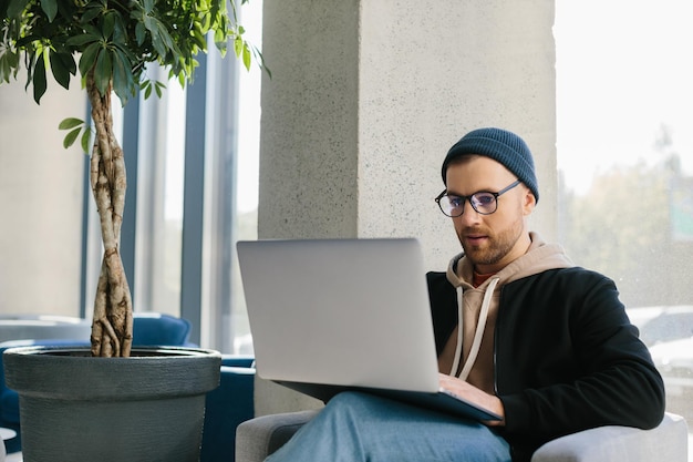 Young handsome freelancer guy working with laptop in office space A man in glasses and a hat is holding a laptop while sitting in a chair in the hall