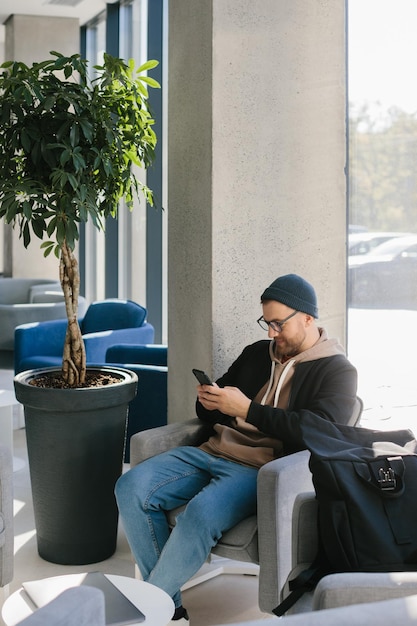 A young handsome guy with a phone in his hands is waiting for a job interview in an office space A man in glasses and a cap is holding a phone while sitting in a chair in the hall