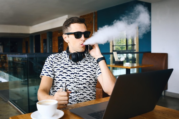 Young handsome hipster man insunglasse sitting in cafe with a cup of coffee vaping