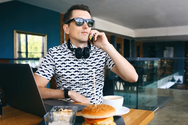 Young handsome hipster man in sunglases sitting in cafe talking mobile phone laptop cup of coffee an