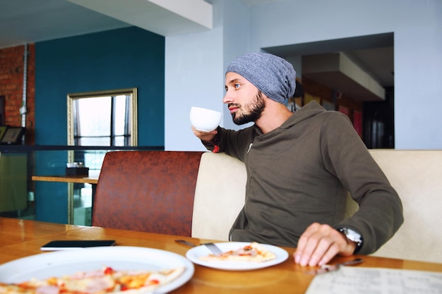 Young handsome hipster man with beard sitting in cafe talking mobile phone holding cup of coffee lap