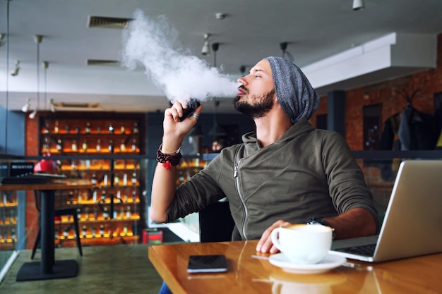 Young handsome hipster man with beard sitting in cafe with a cup of coffee vaping and releases a clo