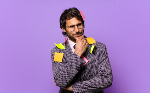 Young handsome indian man smiling with a happy, confident expression with hand on chin, wondering and looking to the side. humorous business concept