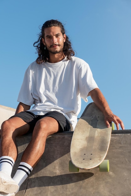 Young handsome male sitting at the top of the skate ramp holding his skateboard He is looking at cam