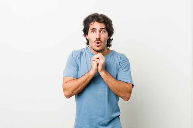 Young handsome man against a white praying for luck, amazed and opening mouth looking to front.