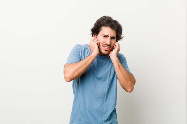 Young handsome man against a white wall covering ears with hands.