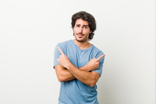 Young handsome man against a white wall points sideways, is trying to choose between two options.