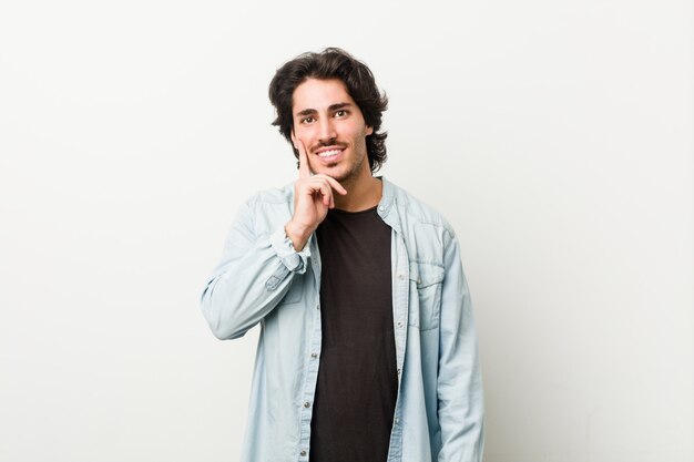 Young handsome man against a white wall smiling happy and confident, touching chin with hand.