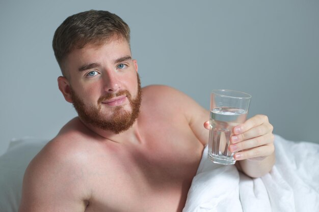 Young handsome man in the bed in bedroom at home in the morning lying under white blanket and drinking pure fresh water from glass