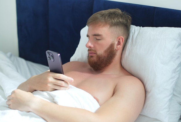 Young handsome man in the bed in bedroom at home in the morning lying under white blanket using cell mobile phone looking at screen of smartphone