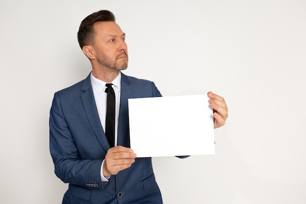 Young handsome man in bussines suit with beard holding empty paper blank in hand on white background, looking to side,