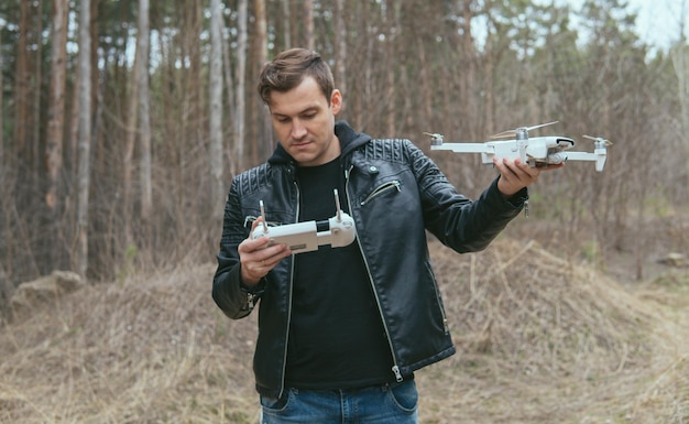 Young handsome man in casual clothes holding white drone and remote control in forest Adult guy going to launch his drone in woodland